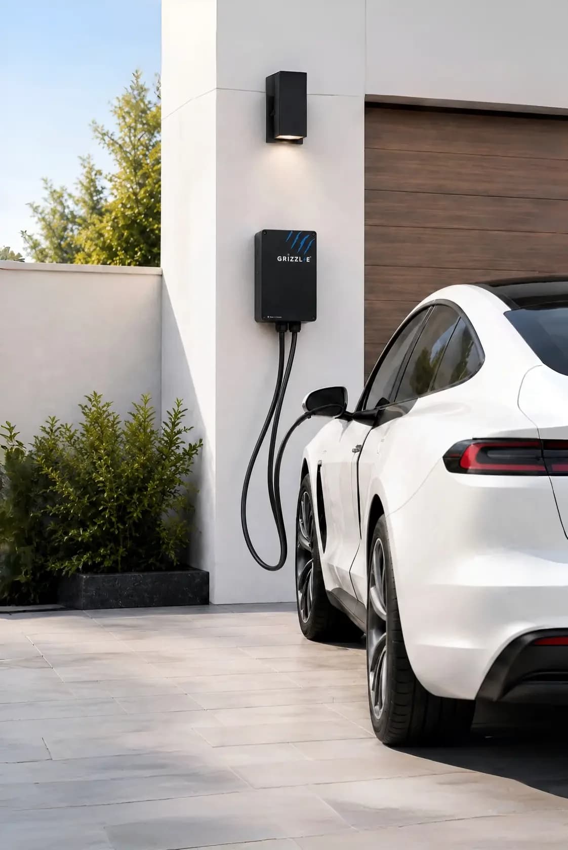 A white electric car charging from a wall-mounted Grizzl-E charger beside a garage